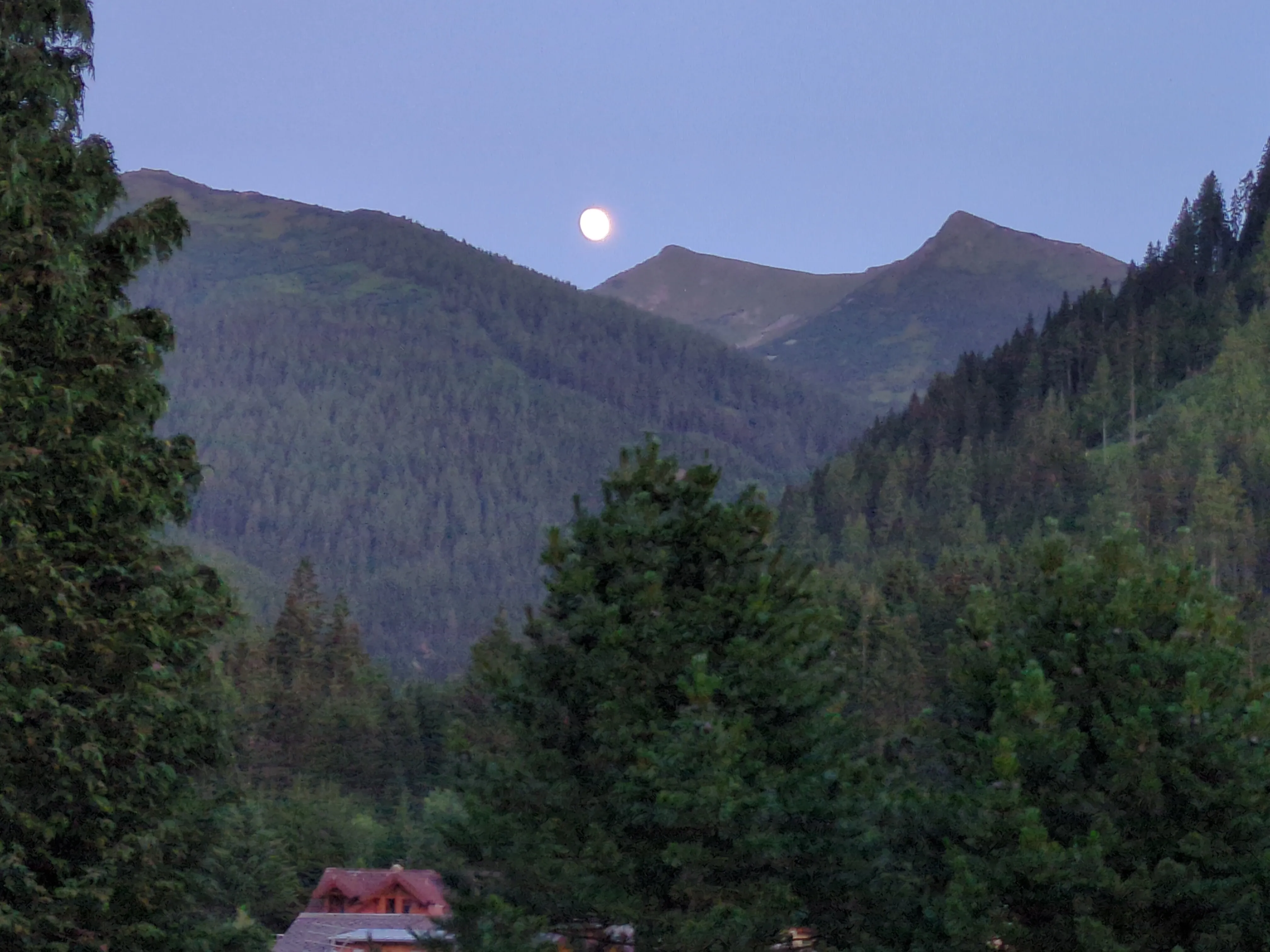 Ein Blick über das von grünen Bäumen verdeckte Dorf hin zu den nahegelegenen Bergen. In der Dämmerung ist der Mond direkt über den Bergen zu sehen.